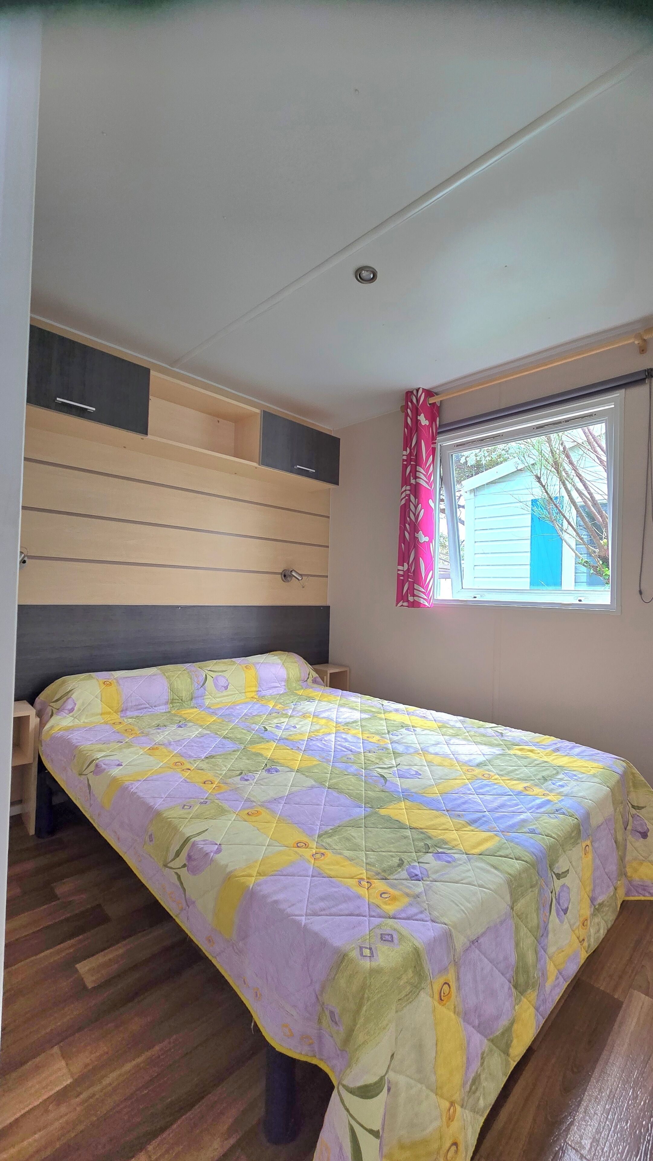 Une chambre à coucher avec un lit, une tête de lit en bois, une fenêtre avec des rideaux et une vue sur un bâtiment à l’extérieur.