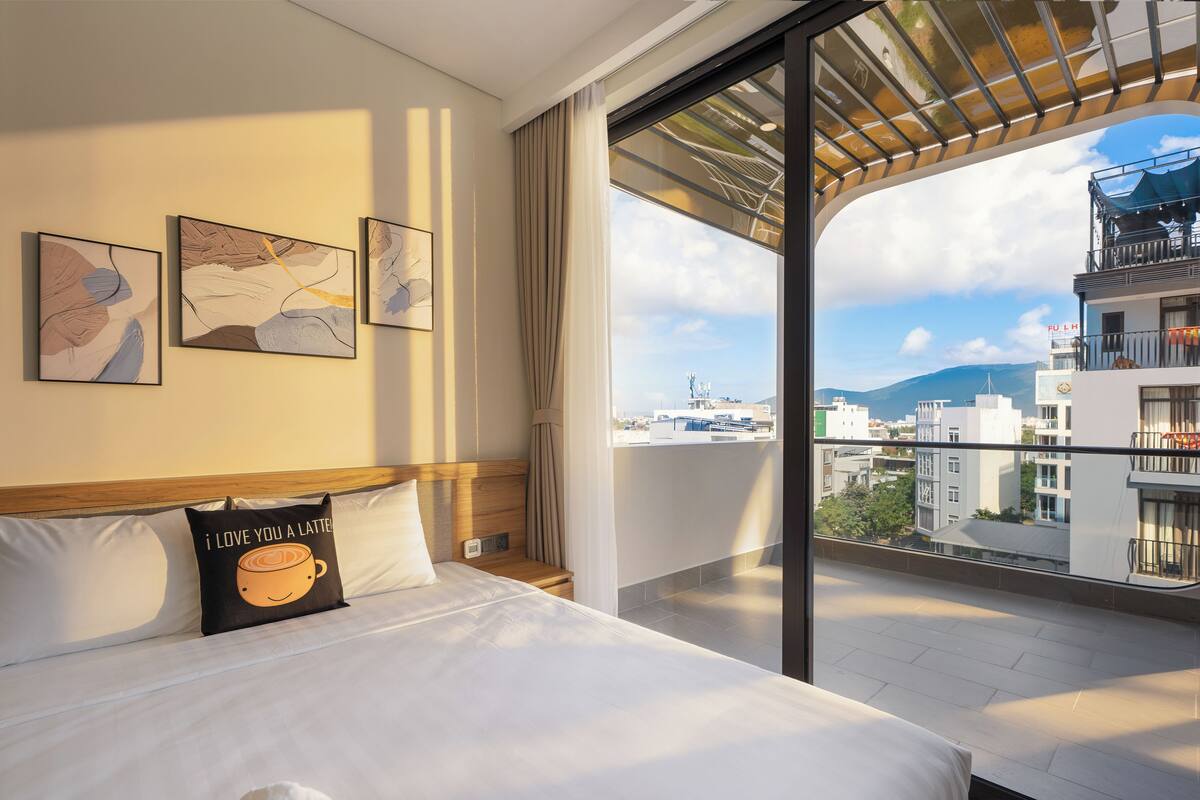 Una habitación de hotel con cama, un almohadón con diseño de taza de café y un amplio ventanal con vistas a edificios y un balcón.