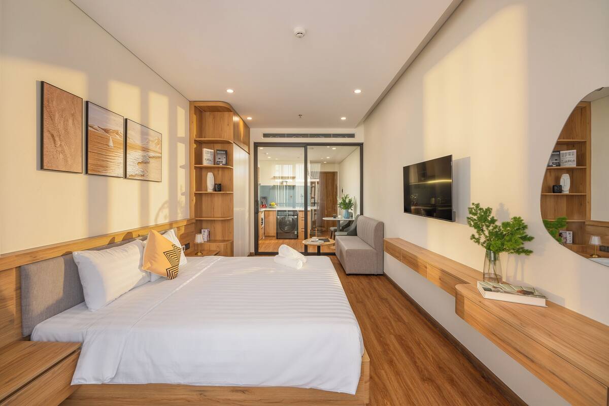 Habitación de hotel moderna con una cama grande, televisor, zona de estar y vistas a la cocina.