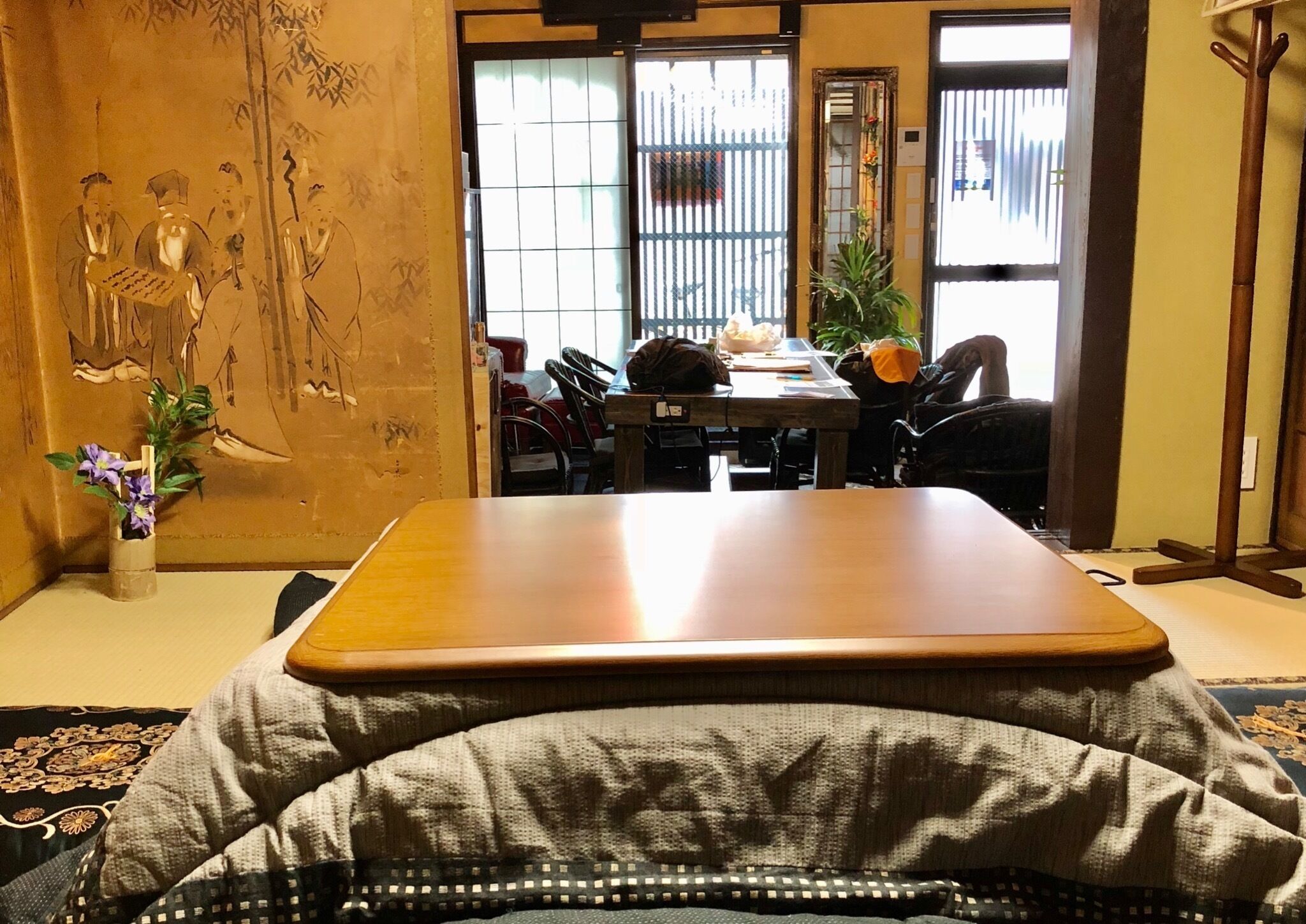 Perinteinen japanilaistyylinen huone, jossa on tatamimatto, matala puupöytä ja näkymä ruokailutilaan, jossa on tuoleja ja kasvi.