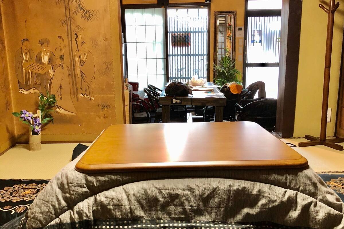 Um quarto em estilo japonês tradicional, com tatames, uma mesa de madeira baixa e vista para uma área de refeições com cadeiras e uma planta.