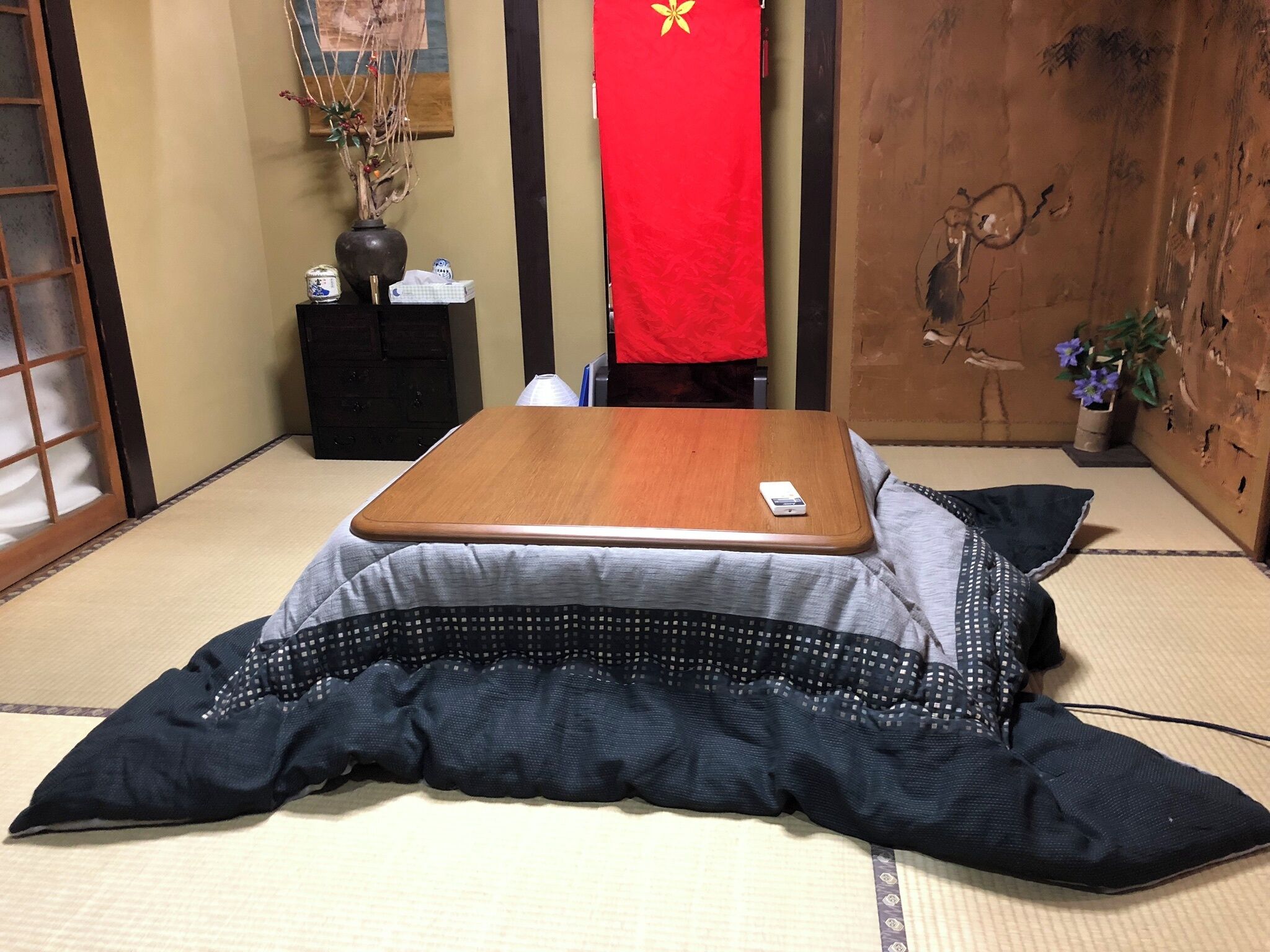 A tatami mat room with a tatami mat bed, a small wooden table, a red hanging object, and a wall with a traditional painting.