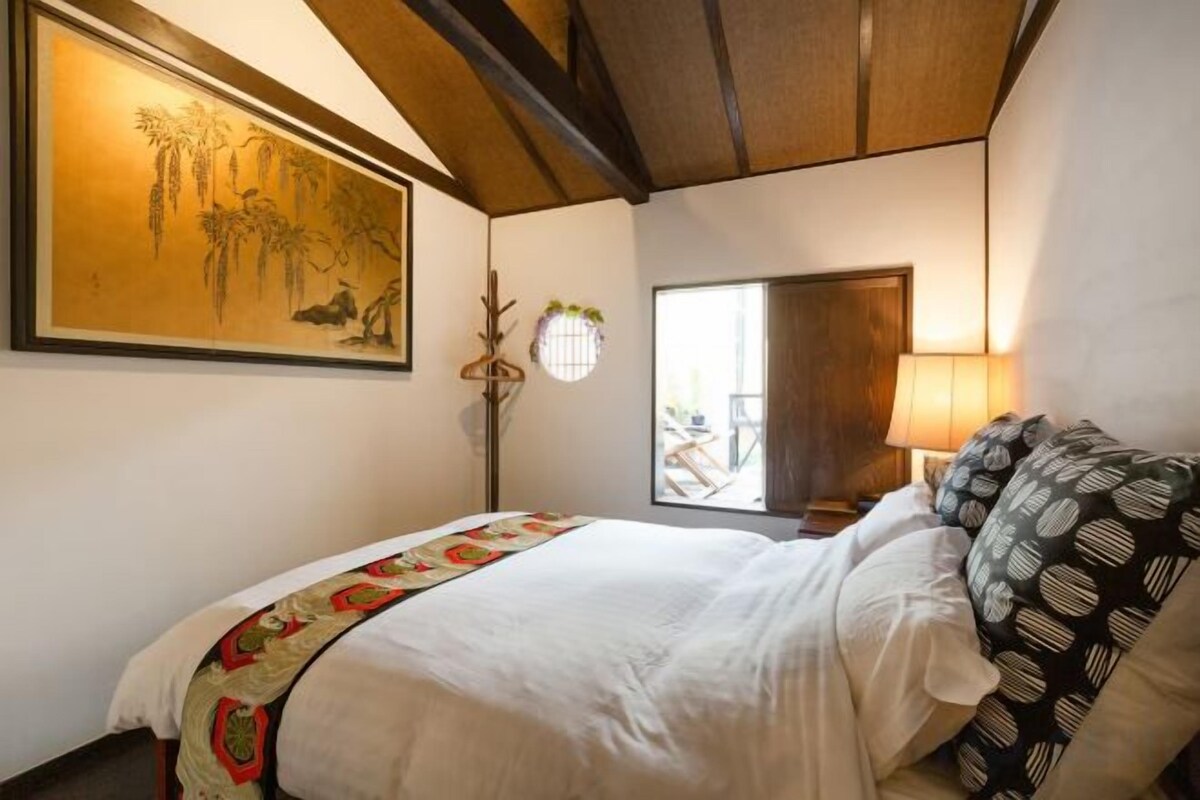 A bedroom with a bed, a nightstand, a lamp, a window, and a painting on the wall.