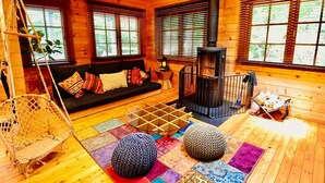 A cozy cabin interior with a wooden floor, a black sofa with colorful cushions, a hanging chair, a fireplace, and a small table with a rug. - Asobi No Cottage Hatsukaichi (Hatsukaichi)