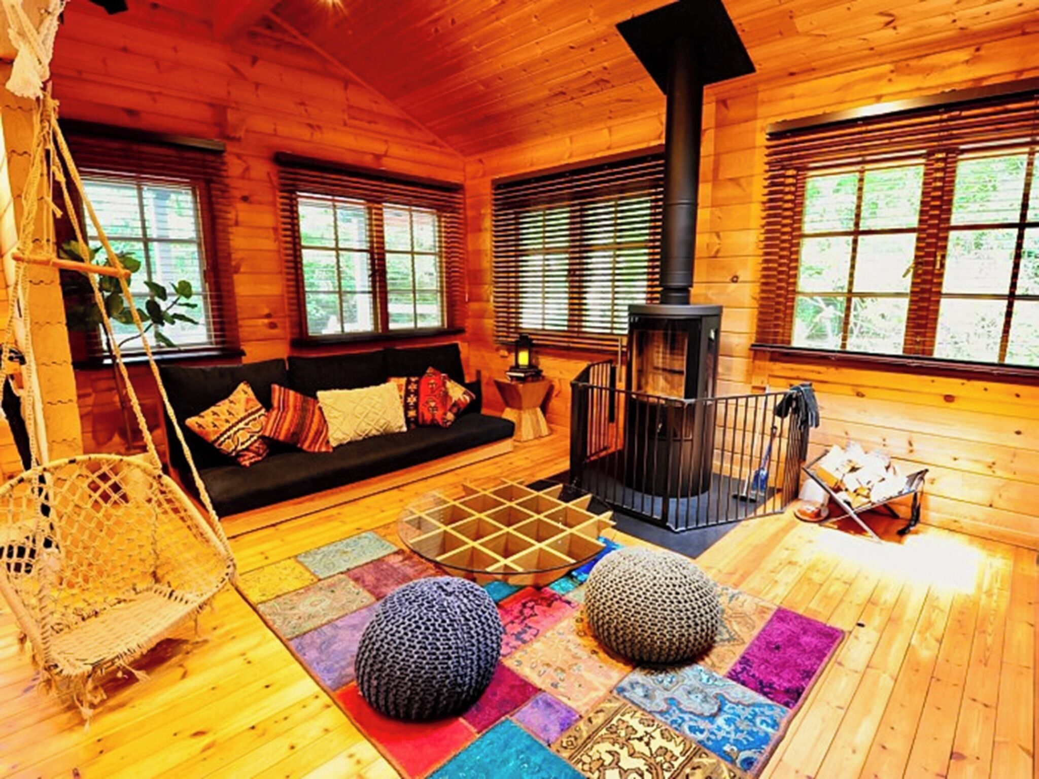 Een gezellig cabin interieur met een houten vloer, een zwarte bank met kleurrijke kussens, een hangstoel, een zwarte open haard en een raam met luxaflex.