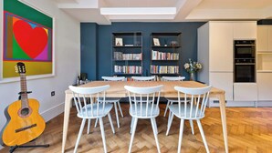A dining area with a wooden table and white chairs, a bookshelf, and a guitar.