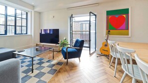 A modern living room with a flat-screen TV, a blue armchair, a wooden dining table with white chairs, a guitar, and a large window.