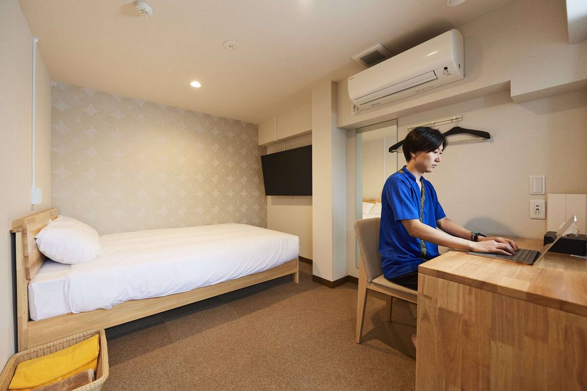 A room with a bed, a desk with a laptop, an air conditioner, and a television.