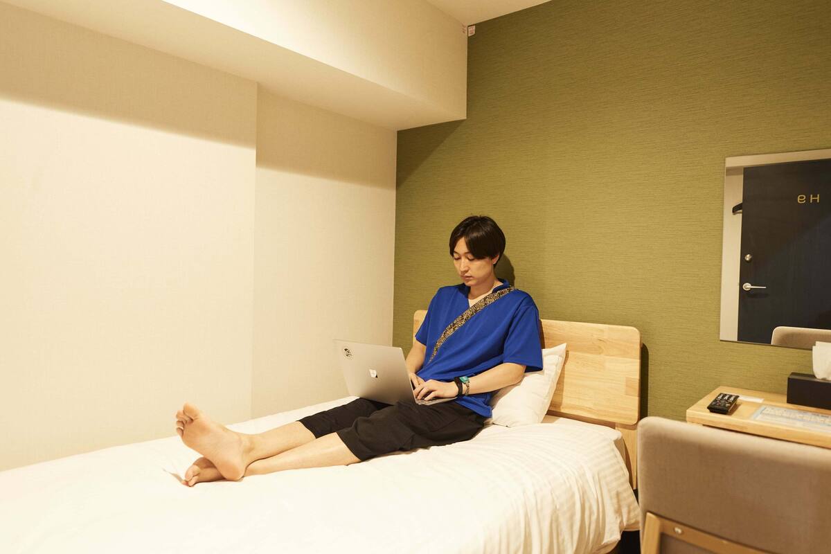 A person sitting on a bed in a hotel room with a laptop, a bedside table with a remote control, and a door with a handle.