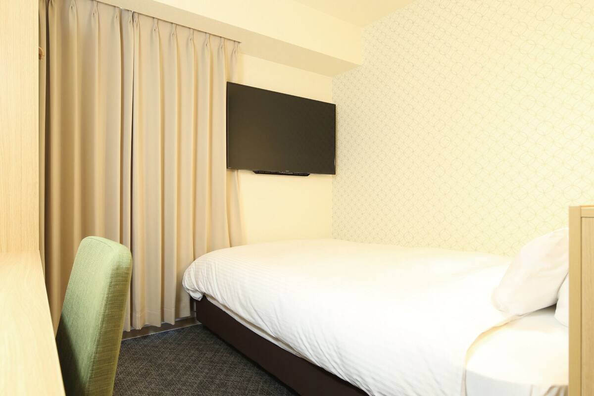 A hotel room with a bed, a chair, a TV mounted on the wall, and a window with curtains.