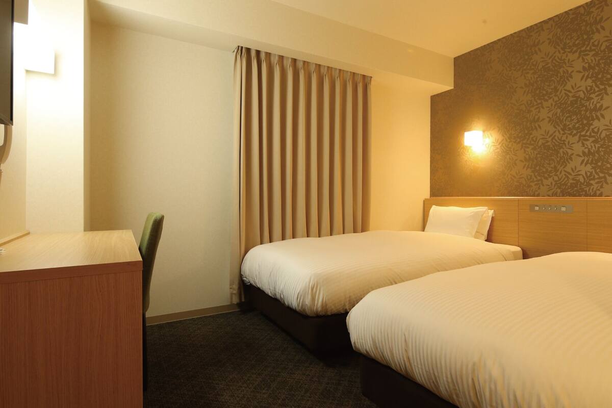 A hotel room with two beds, a desk, a chair, and a wall-mounted light fixture.