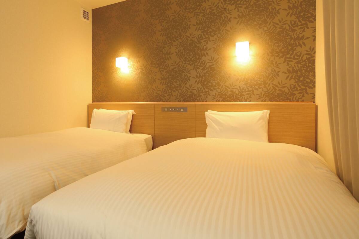 A hotel room with two single beds, a headboard with a control panel, and two wall-mounted lights.