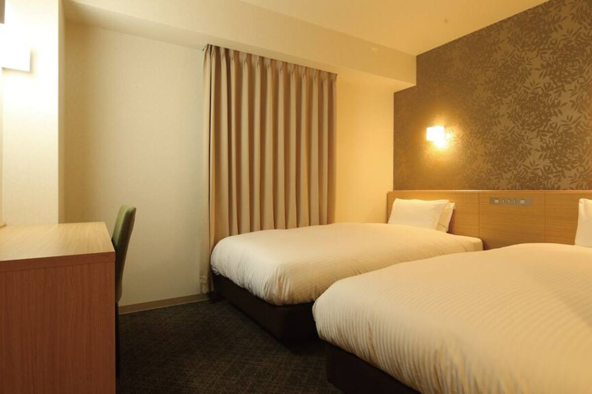 A hotel room with two beds, a desk, a chair, and a wall-mounted light fixture.