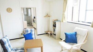 Een kamer met een bank, een stoel, een klein tafeltje, een klok, een raam en een ventilator.