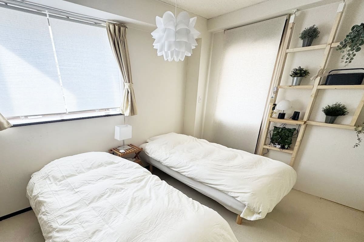 Un dormitorio con dos camas, una ventana con cortinas, una mesita de noche con lámpara y una estantería con plantas.