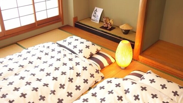 Een traditionele Japanse kamer met tatami vloerbedekking, twee bedden met kruispatroon beddengoed, een shoji schuifwand en een klein plankje met decoratieve objecten.