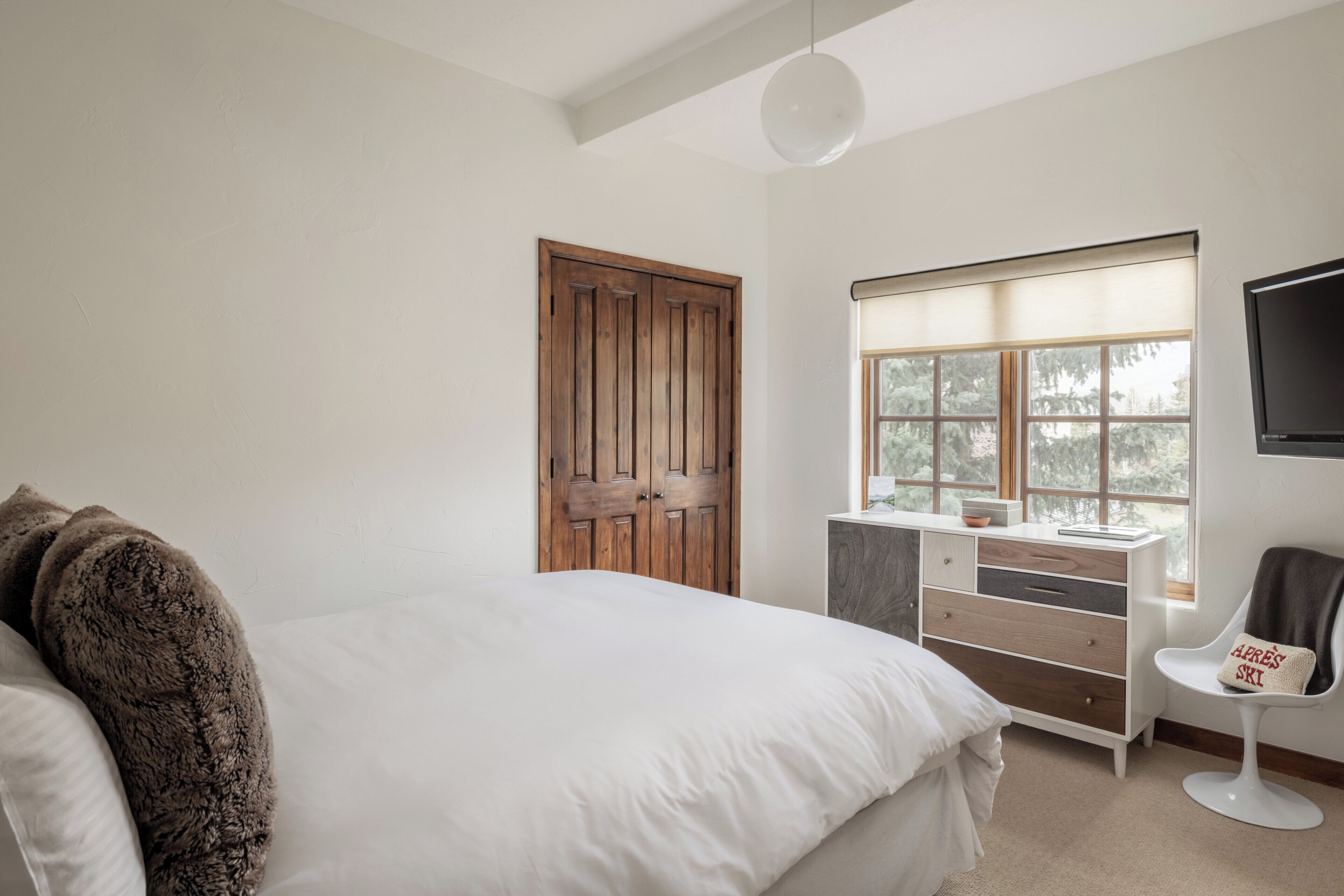 A bedroom with a large bed, a wooden door, a window with blinds, a white dresser, and a TV mounted on the wall.