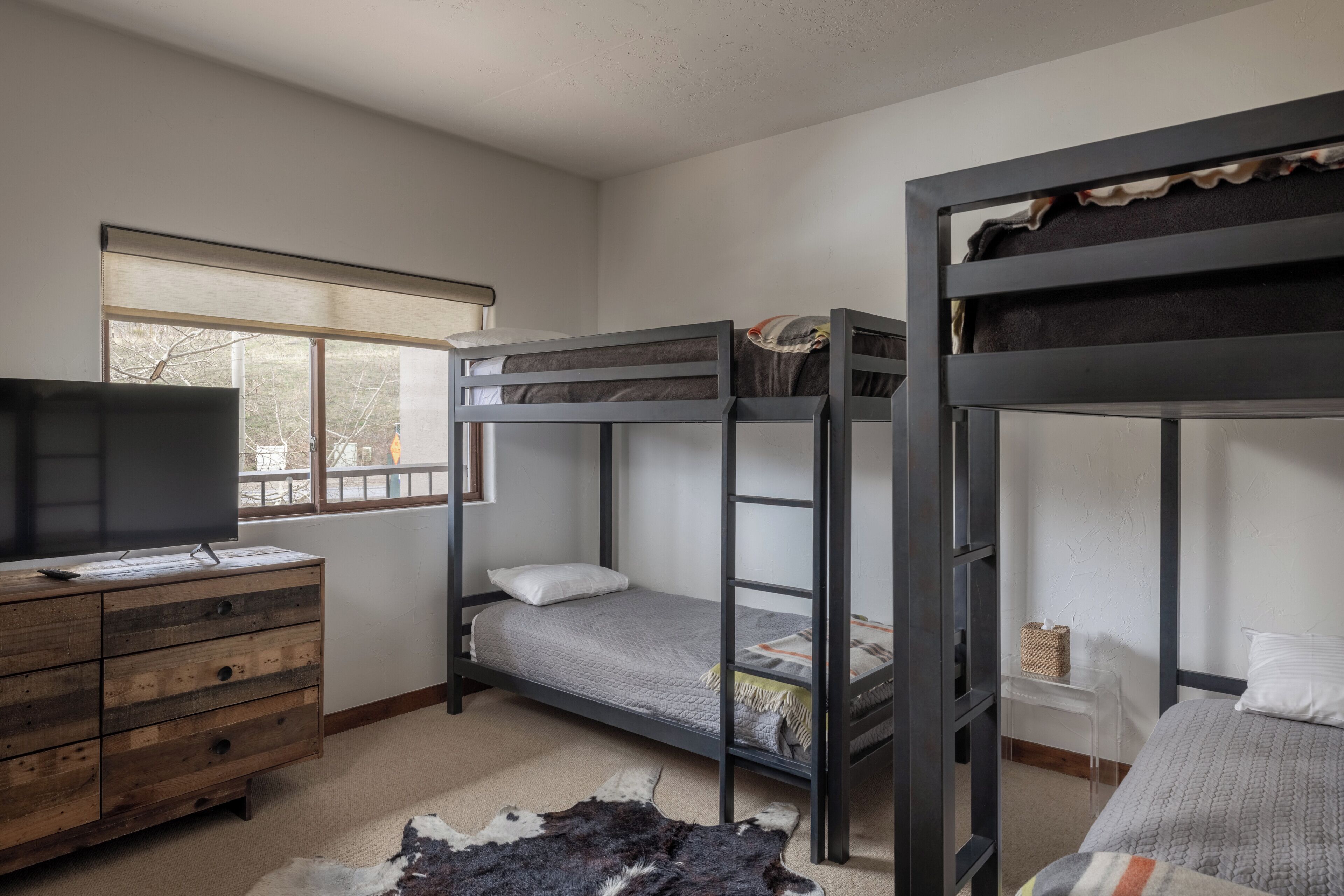 A bunk bed room with a TV on a wooden dresser, a window with a view, and a rug.
