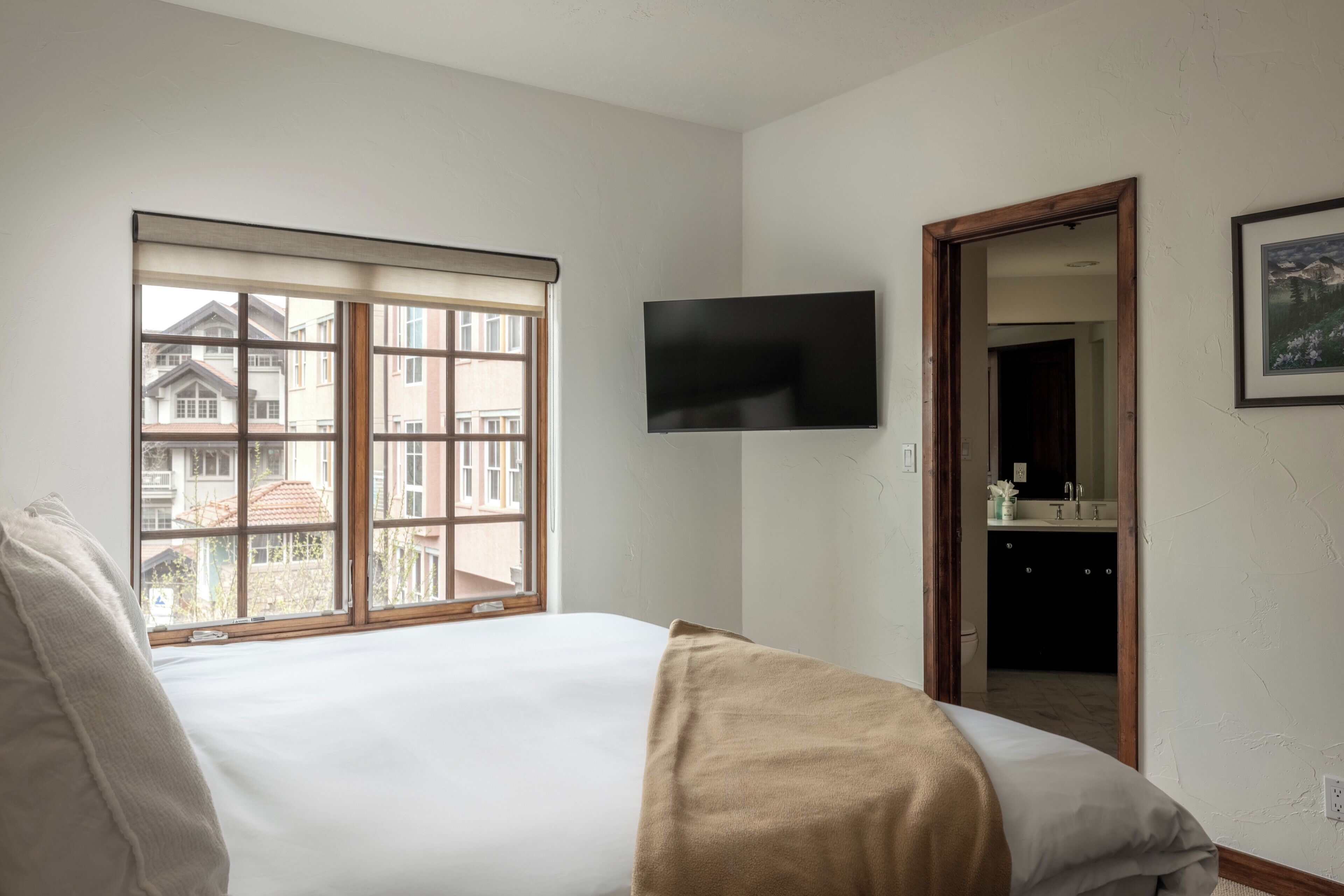 A bedroom with a bed, a TV mounted on the wall, a window with a view of buildings, and a door leading to a bathroom.