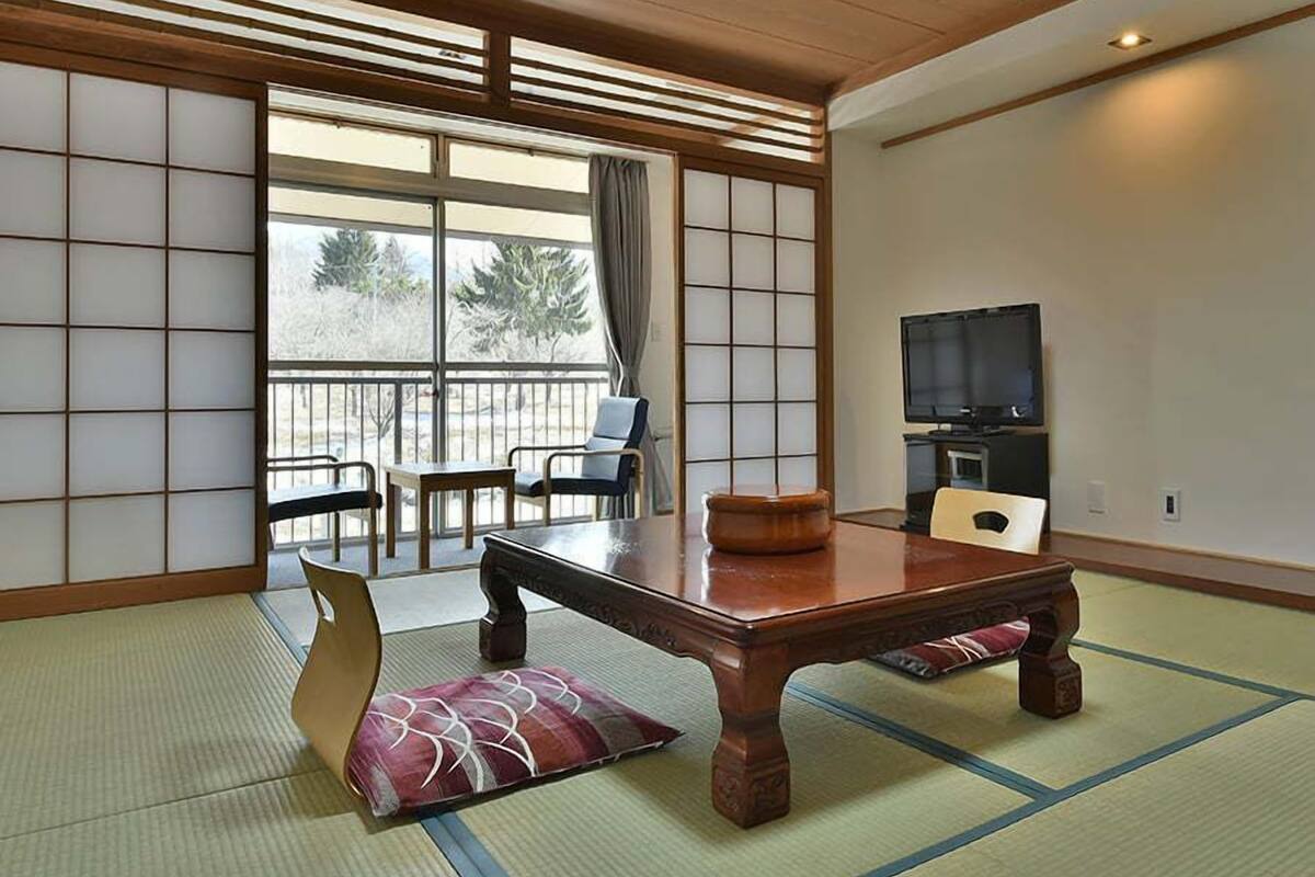 A traditional Japanese-style room with tatami flooring, a low wooden table, a television, and a view of the outdoors.