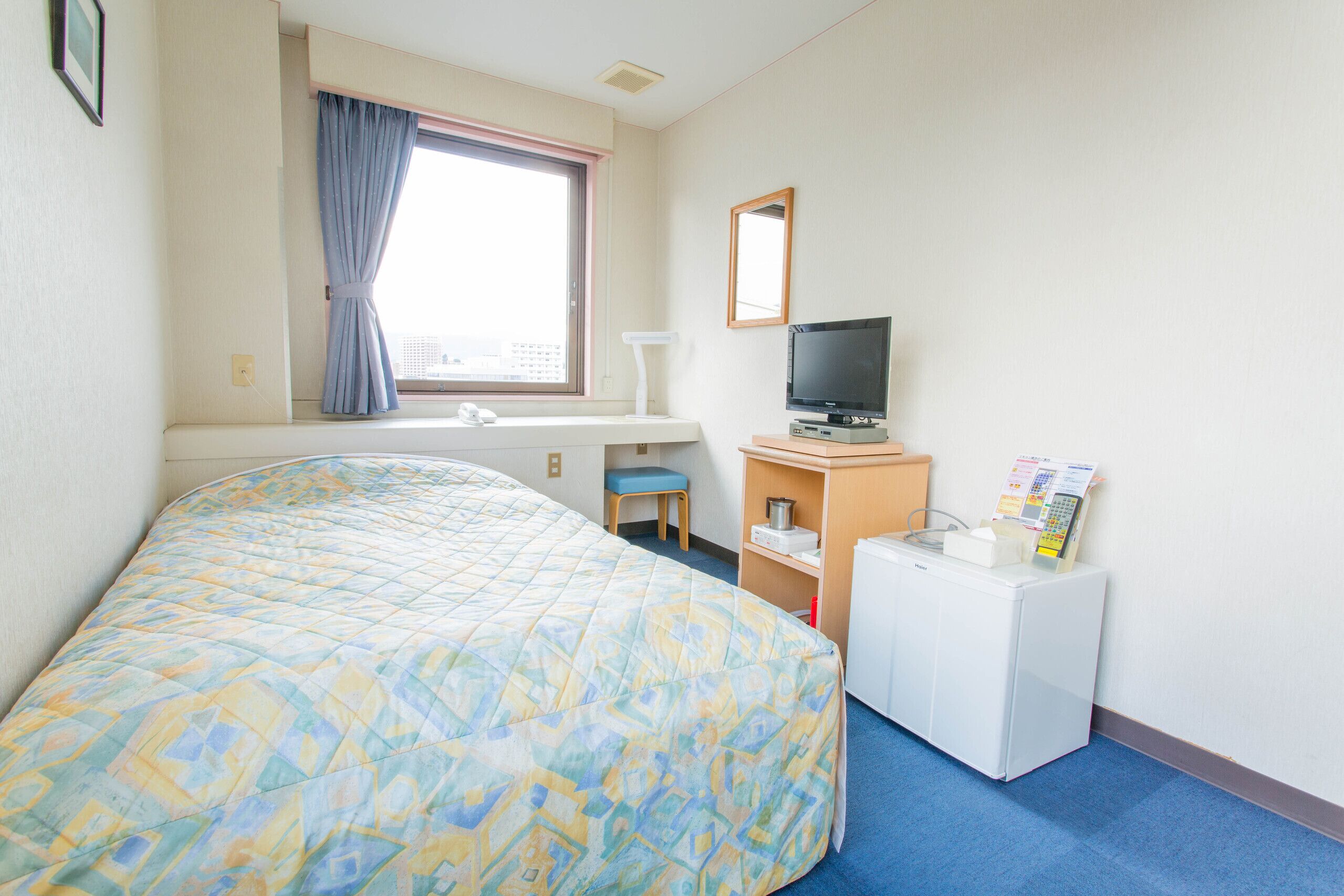 A hotel room with a bed, a desk with a television, a small refrigerator, and a window with curtains.