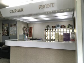 Zona de recepción de un hotel con un mostrador blanco, una caja registradora, un reloj y varios carteles informativos en la pared.