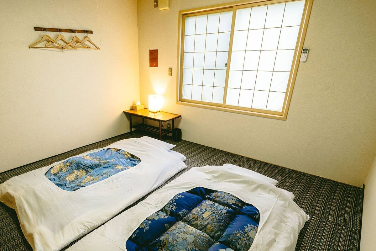 A hotel room with two beds, a bedside table with a lamp, a window with a view, and a wall-mounted shelf with hangers.