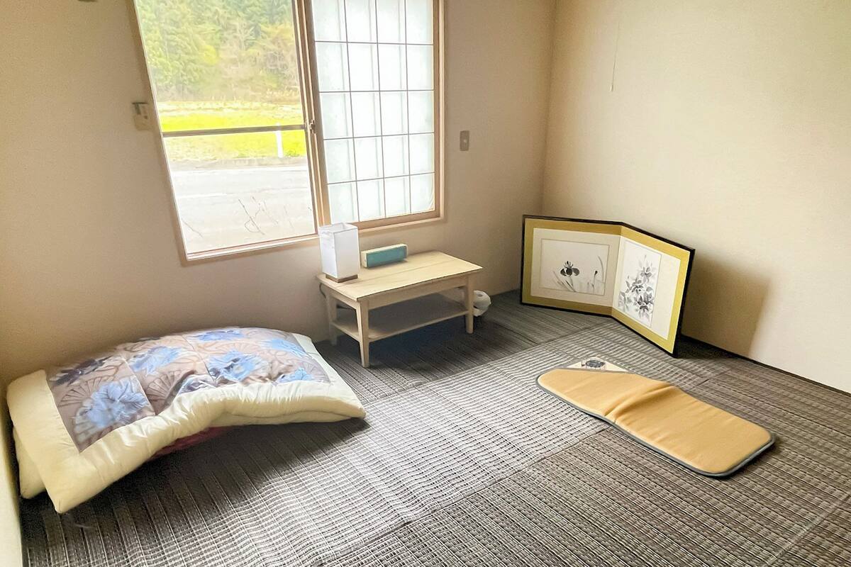 A small room with a bed, a desk, a chair, a window, and a mat on the floor.