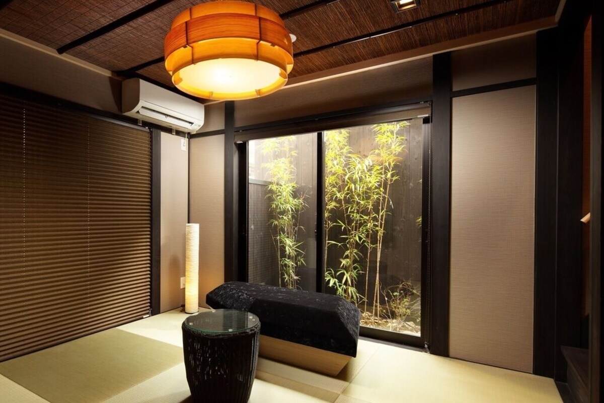 A traditional Japanese room with tatami flooring, a sliding door, a view of bamboo outside, and a modern hanging light fixture.