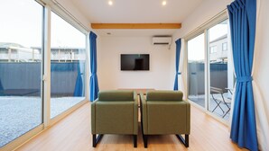 Ein Zimmer mit Fernseher, zwei grünen Stühlen, Holzboden und großen Fenstern mit Vorhängen.