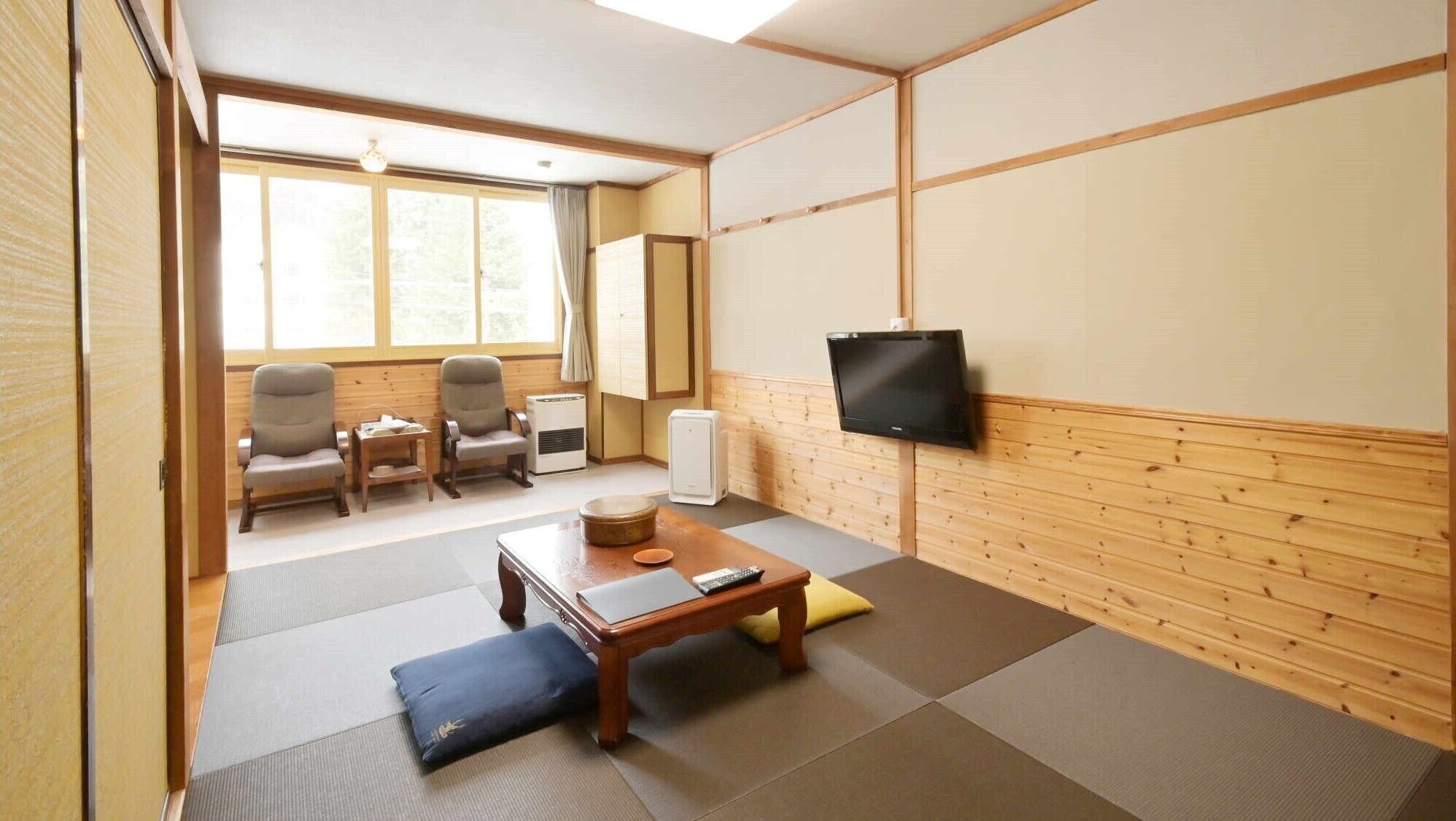 Ein Zimmer mit Tatami-Matten, einem Fernseher, einem Tisch inklusive Fernbedienung und einem Fenster mit Vorhängen.