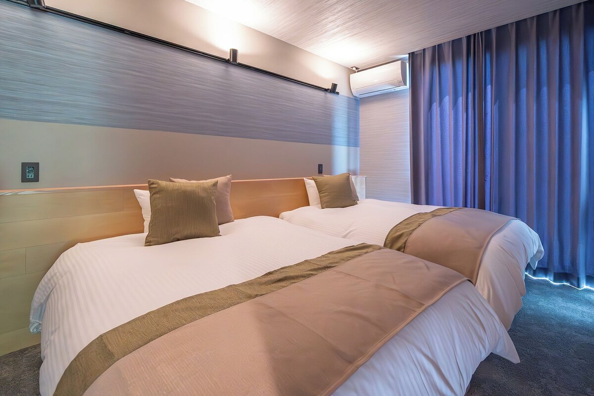 A hotel room with two beds, a headboard, a window with curtains, and an air conditioning unit.