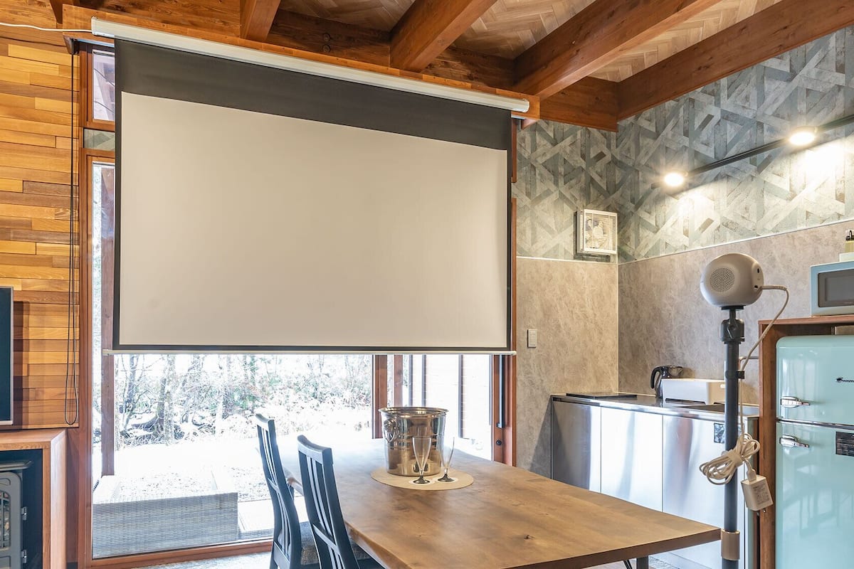 A wooden dining table with chairs, a projector screen, a fireplace, and a kitchen area with a refrigerator and microwave.