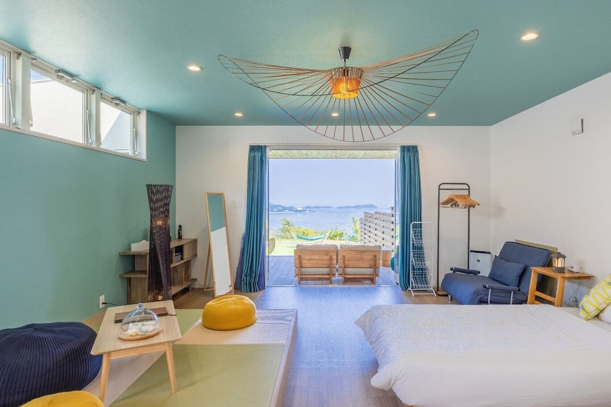 Quarto de hotel com cama, escrivaninha, cadeira, luminária pendente e vista para o mar.