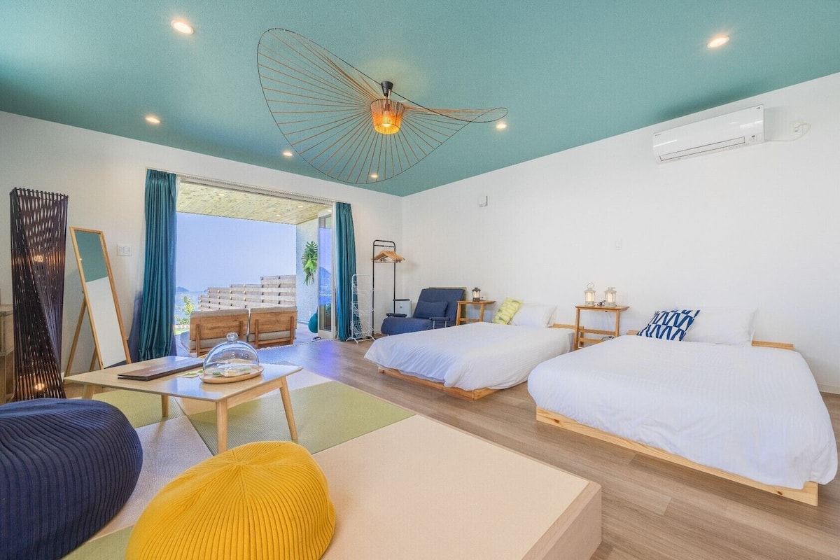 Quarto de hotel com duas camas, uma escrivaninha, uma cadeira, um sofá e uma janela grande com cortinas.