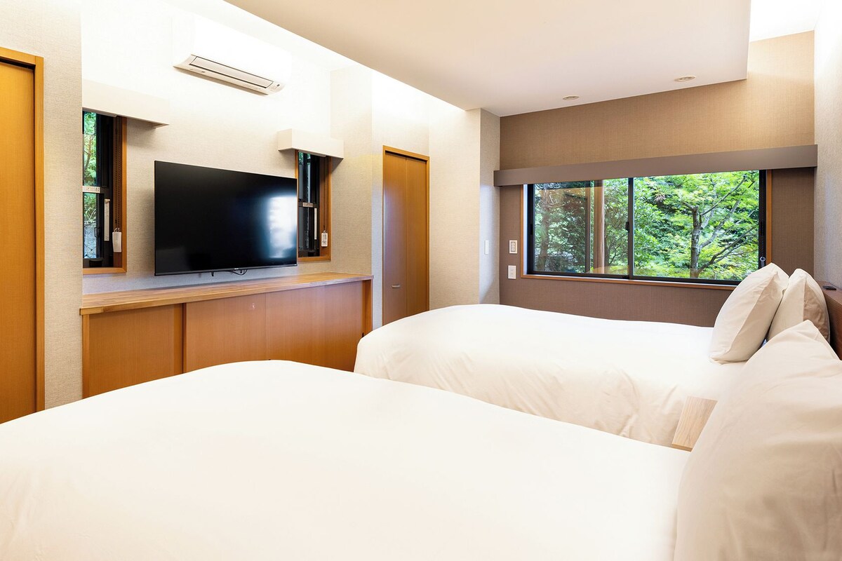A hotel room with two beds, a television, and a window with a view of trees.