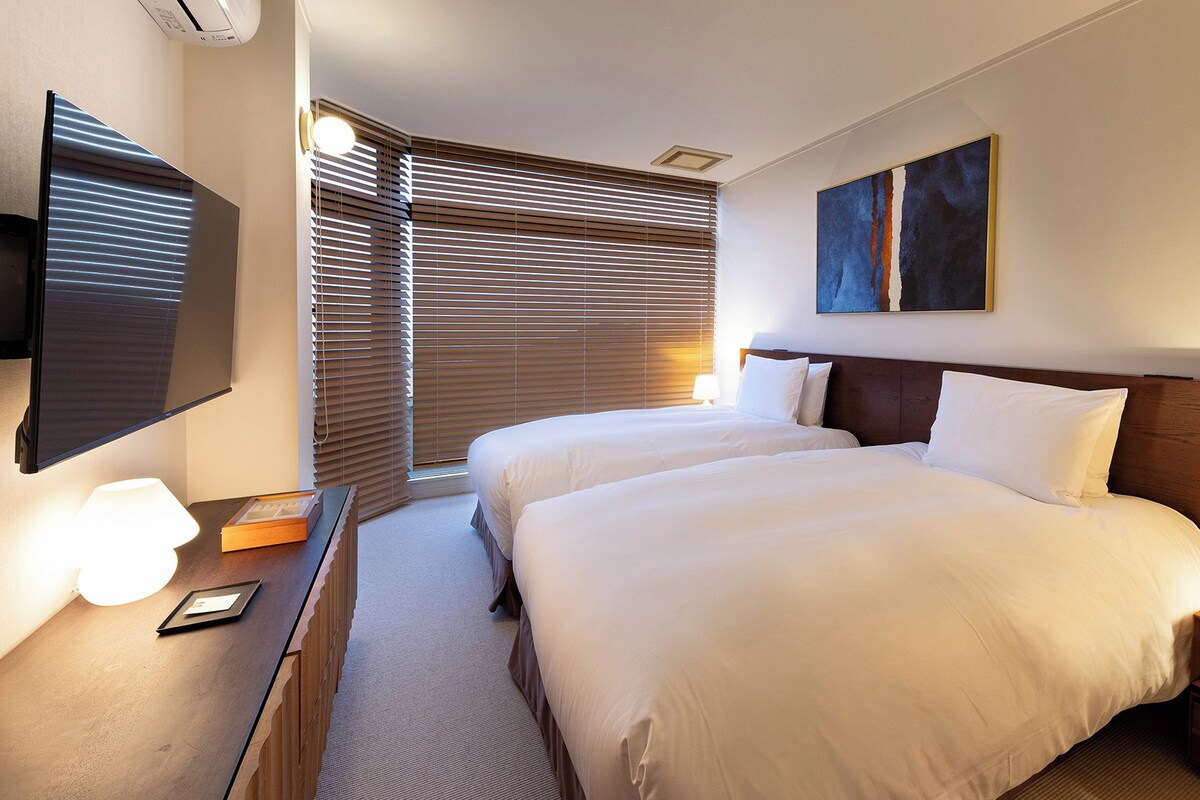 A hotel room with two beds, a flat-screen TV, a painting, and a lamp on the bedside table.