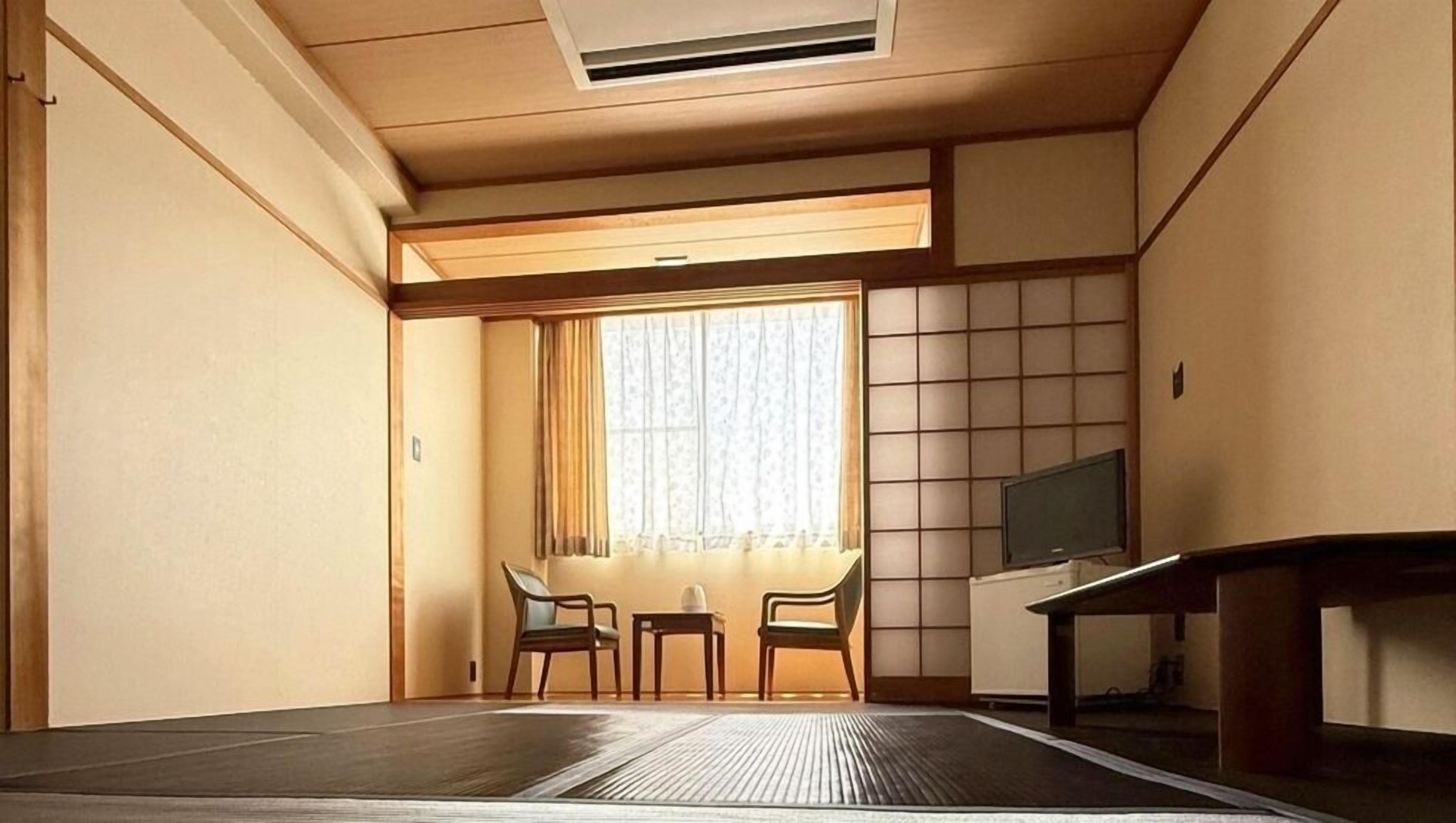 Una habitación con suelo de tatami, una mesa baja con sillas, un televisor sobre un mueble y un biombo con una ventana.