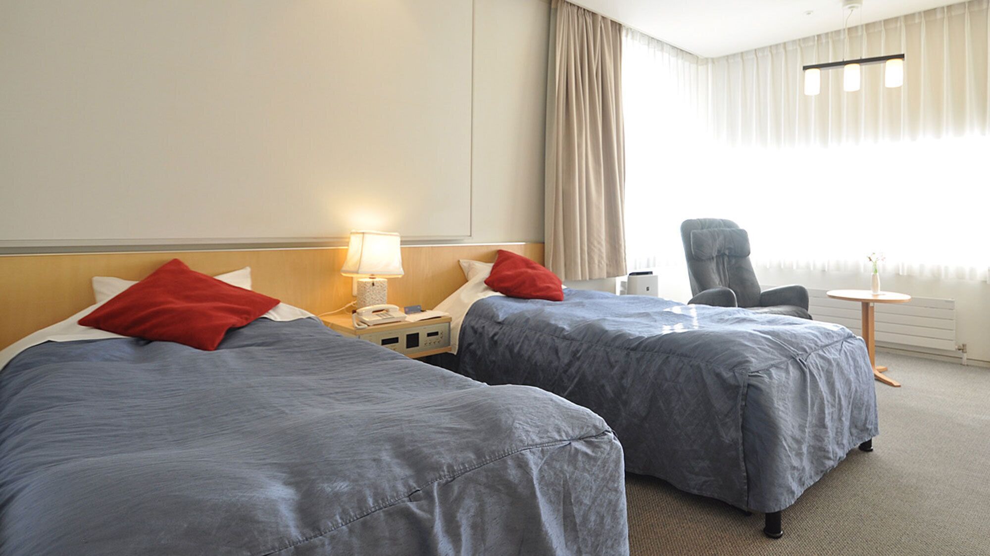 A hotel room with two beds, a chair, a lamp, and a small table.