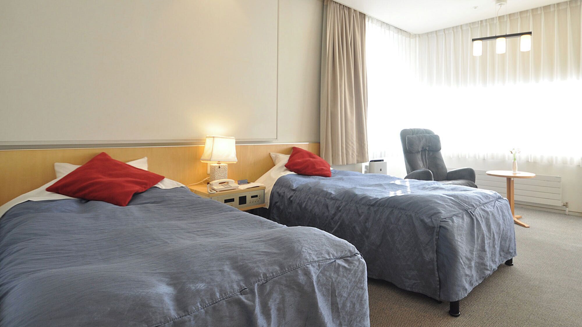 A hotel room with two beds, a chair, a small table, and a lamp.