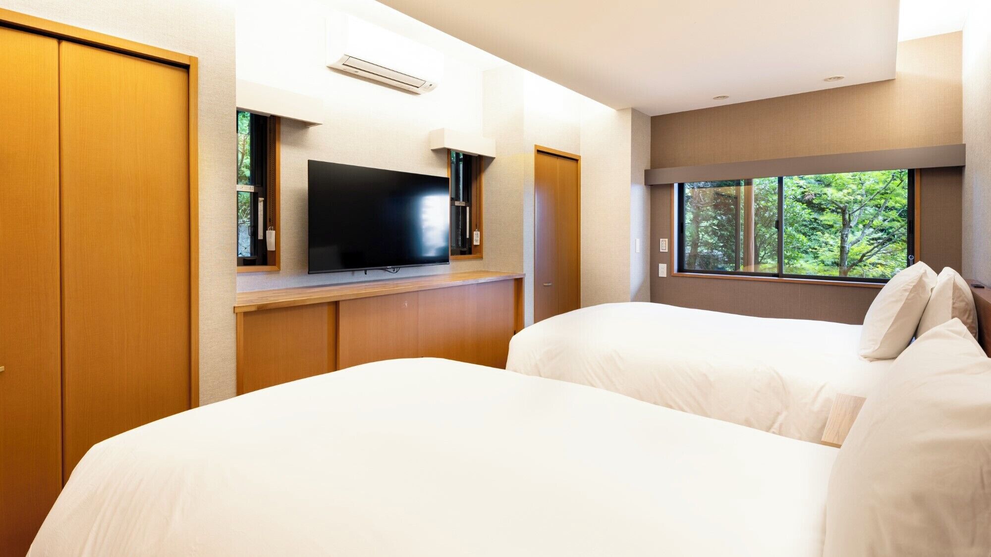 Ein Hotelzimmer mit zwei Betten, einem Fernseher auf einem Holzboden und einem Fenster mit Blick auf Bäume.