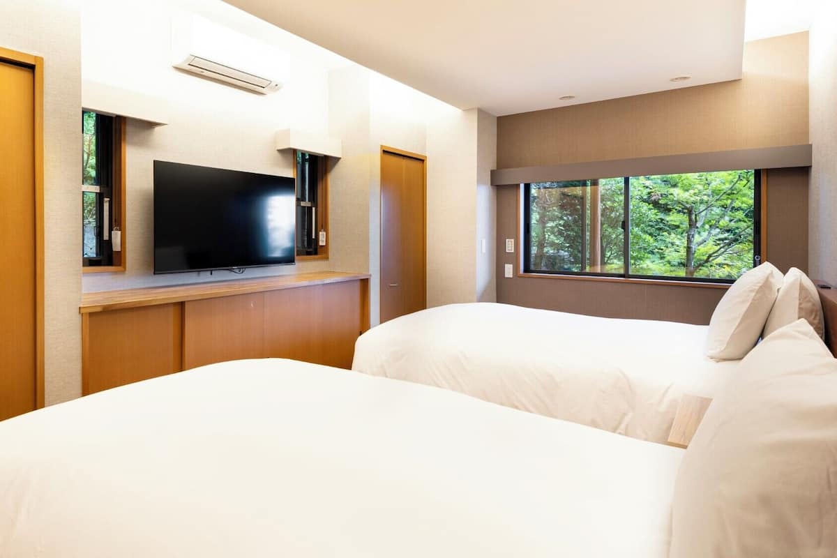 A hotel room with two beds, a TV on a wooden shelf, and a window with a view of trees.
