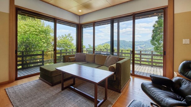 A room with a wooden table, green sofa, and black leather chair, large windows with a view of greenery and hills. - Glampetit Koh (Atami)