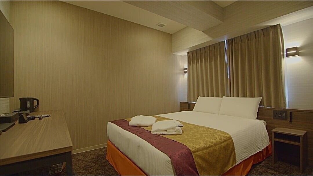 A hotel room with a bed, a desk, a nightstand, a coffee maker, and a window with curtains.