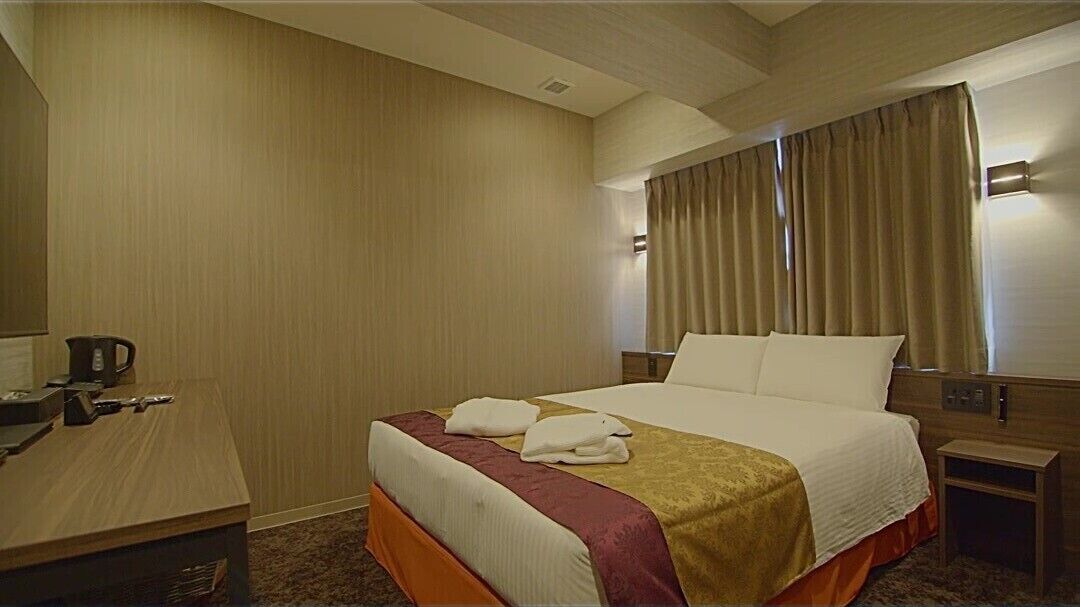 Ein Hotelzimmer mit Bett, Schreibtisch, Nachttisch, Lampe und einem Fenster mit Vorhängen.