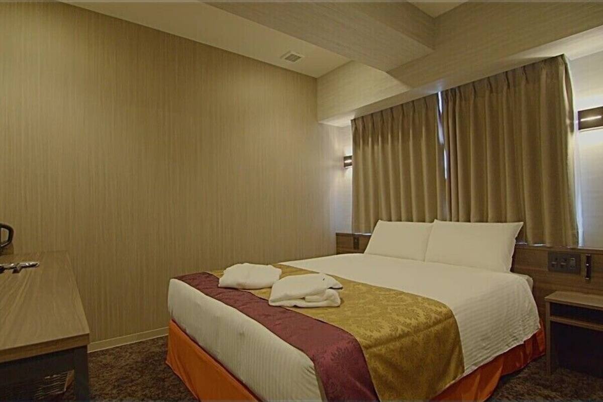 Una habitación de hotel con cama, escritorio, mesita de noche, lámpara y ventana con cortinas.