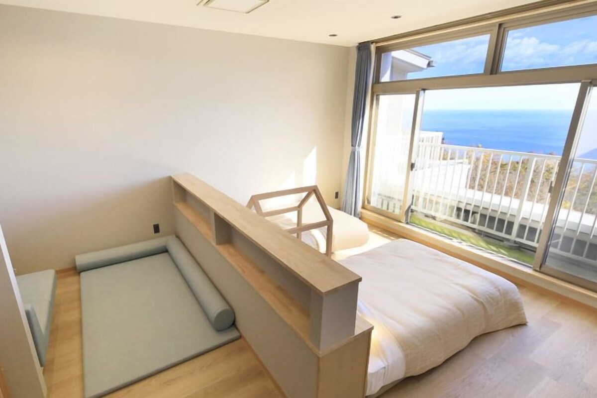 A modern bedroom with a bed, a small seating area, and a large window offering a view of the sea.