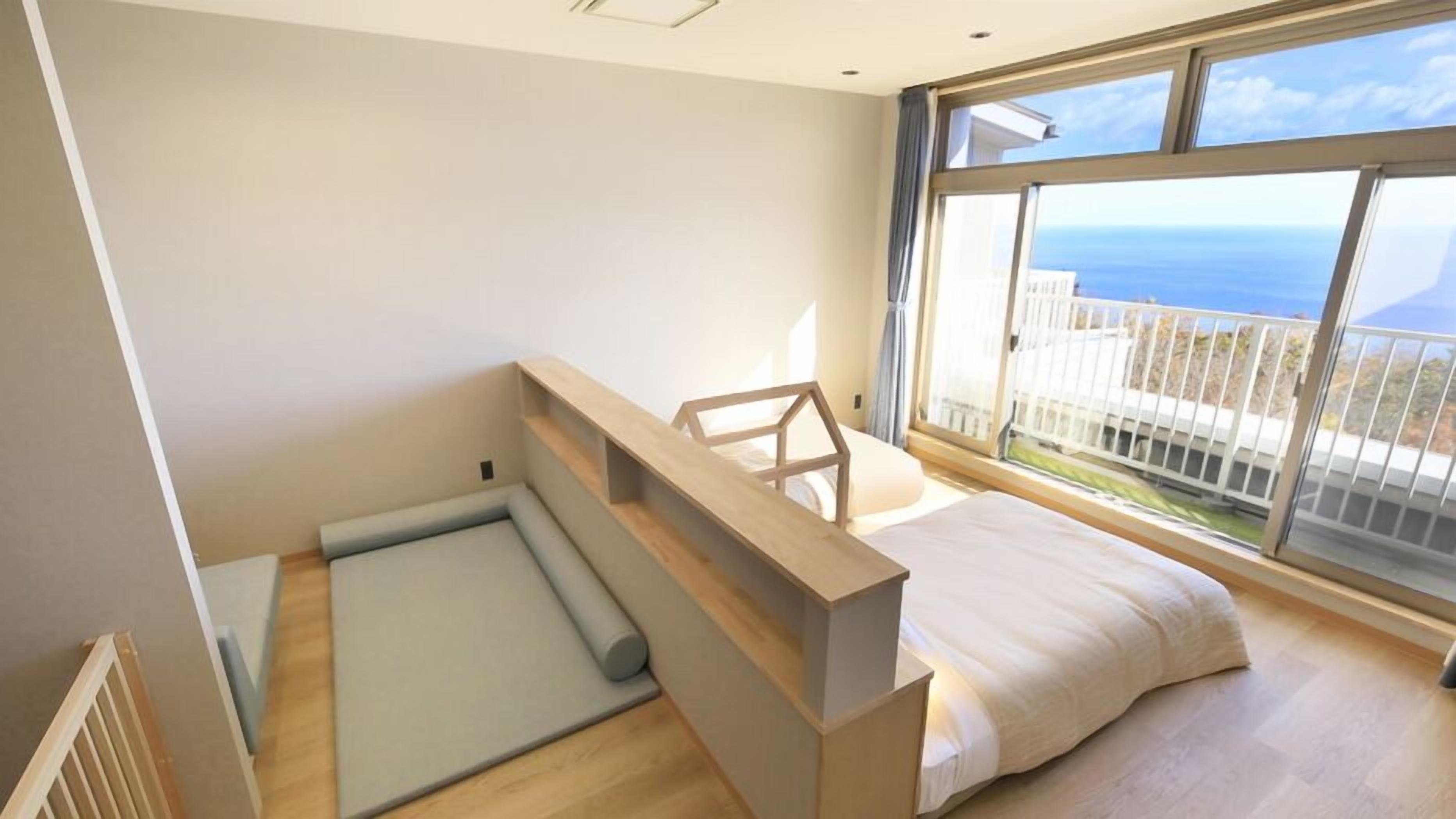 A modern bedroom with a bed, a small seating area, and a large window offering a view of the sea.