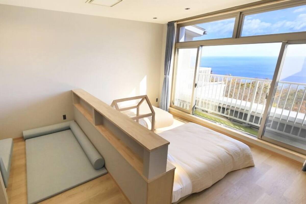 Un dormitorio moderno con cama, un pequeño rincón de descanso y un amplio ventanal con vistas al mar.