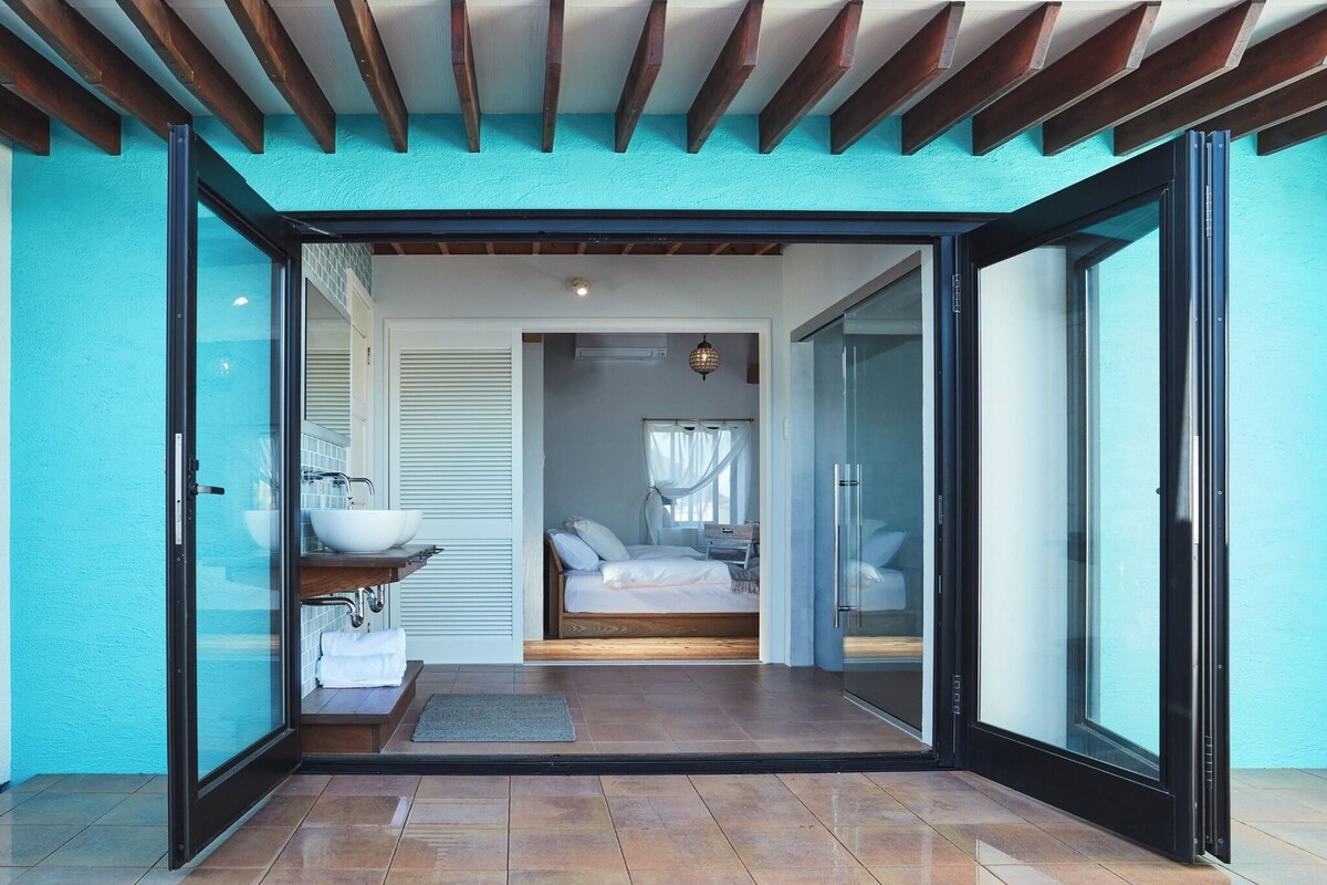 A modern bedroom with a glass door leading to a bathroom, a bed with white linens, and a bedside table.